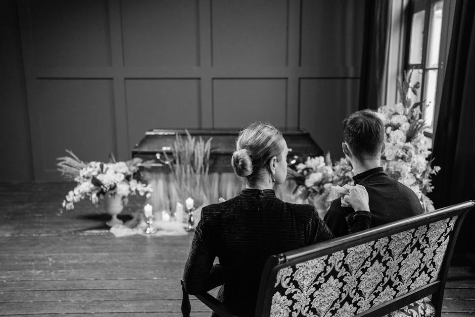 A woman with blonde hair styled in a bun, dressed in a dark textured jacket, sits on an ornate patterned chair holding a small bouquet of light pink tulips. She is facing away from the camera in a sof