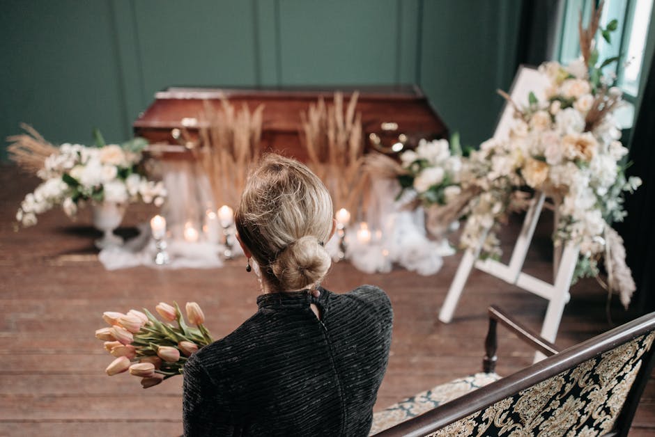 A floral tribute consisting of white lilies with open blooms revealing orange- and brown-streaked stamens, surrounded by closed buds and lush green leaves, rests on a grey stone memorial. In the backg