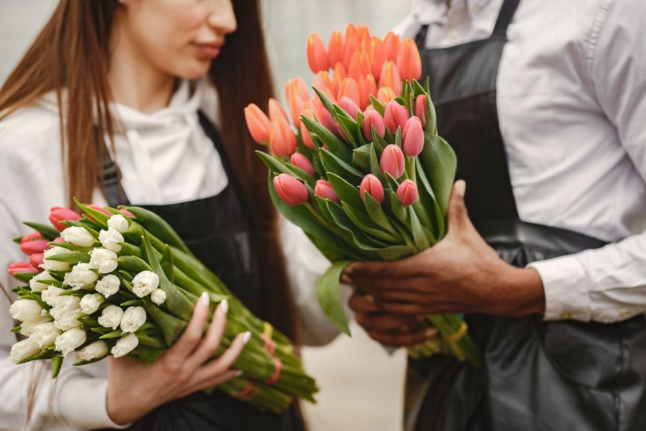 A bouquet of large, fully opened pink peonies with ruffled petals, arranged in a glass vase. The flowers have soft, pastel pink hues with darker pink streaks, and are complemented by lush green leaves