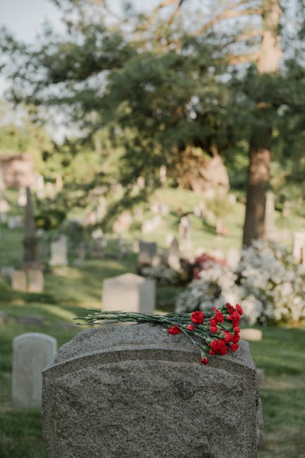 Close-up of an individual's hands arranging a floral tribute on a memorial table. The arrangement includes a bouquet of mixed flowers such as pink roses, white daisies, and purple blooms, wrapped in b
