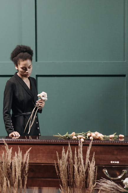 A woman dressed in a black, textured long-sleeve top stands solemnly behind a funeral flower arrangement. In front of her, on a dark wooden table, there are white and pale pink flowers, including anem